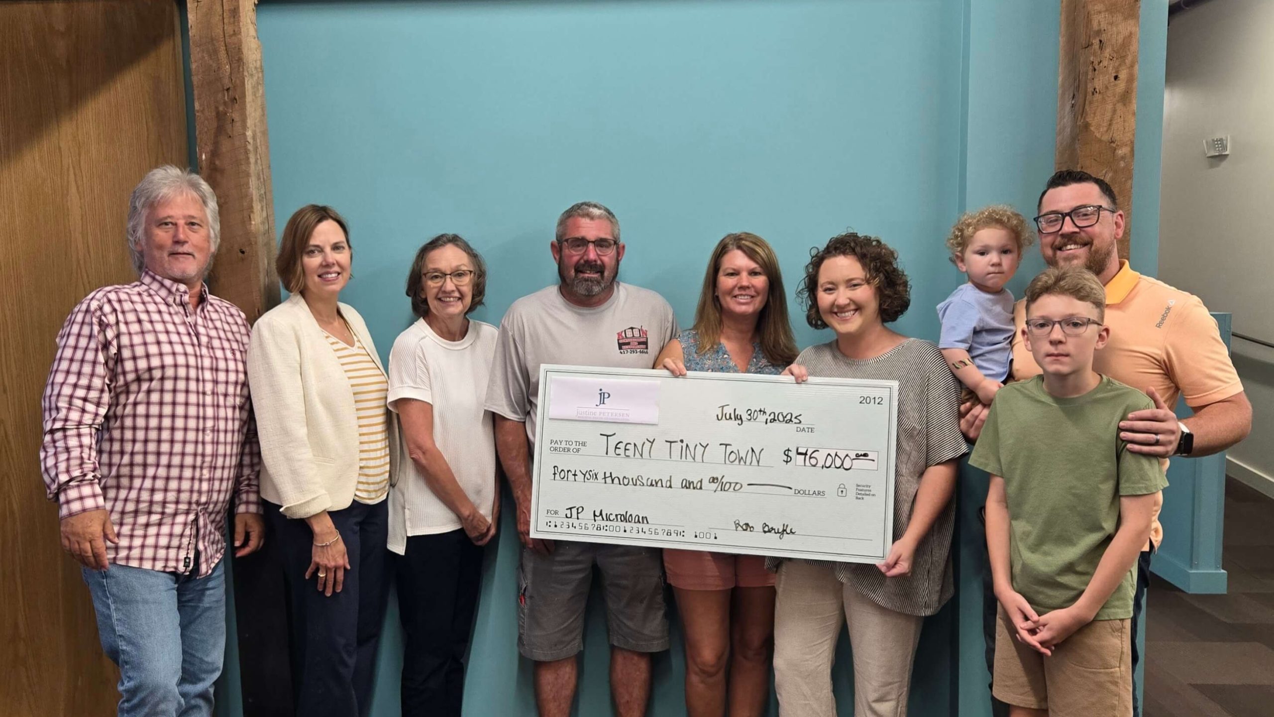 A group of adults and two children standing indoors against a teal wall, smiling and holding an oversized check made out to ‘Teeny Tiny Town’ for $46,000. The group includes men, women, and children gathered closely for a celebratory photo.